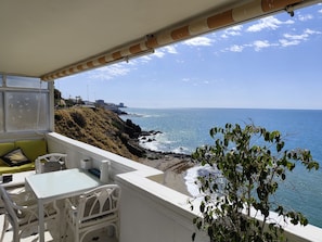 Un balcón con una mesa y sillas blancas, con vista a una zona costera con terreno rocoso y el mar.