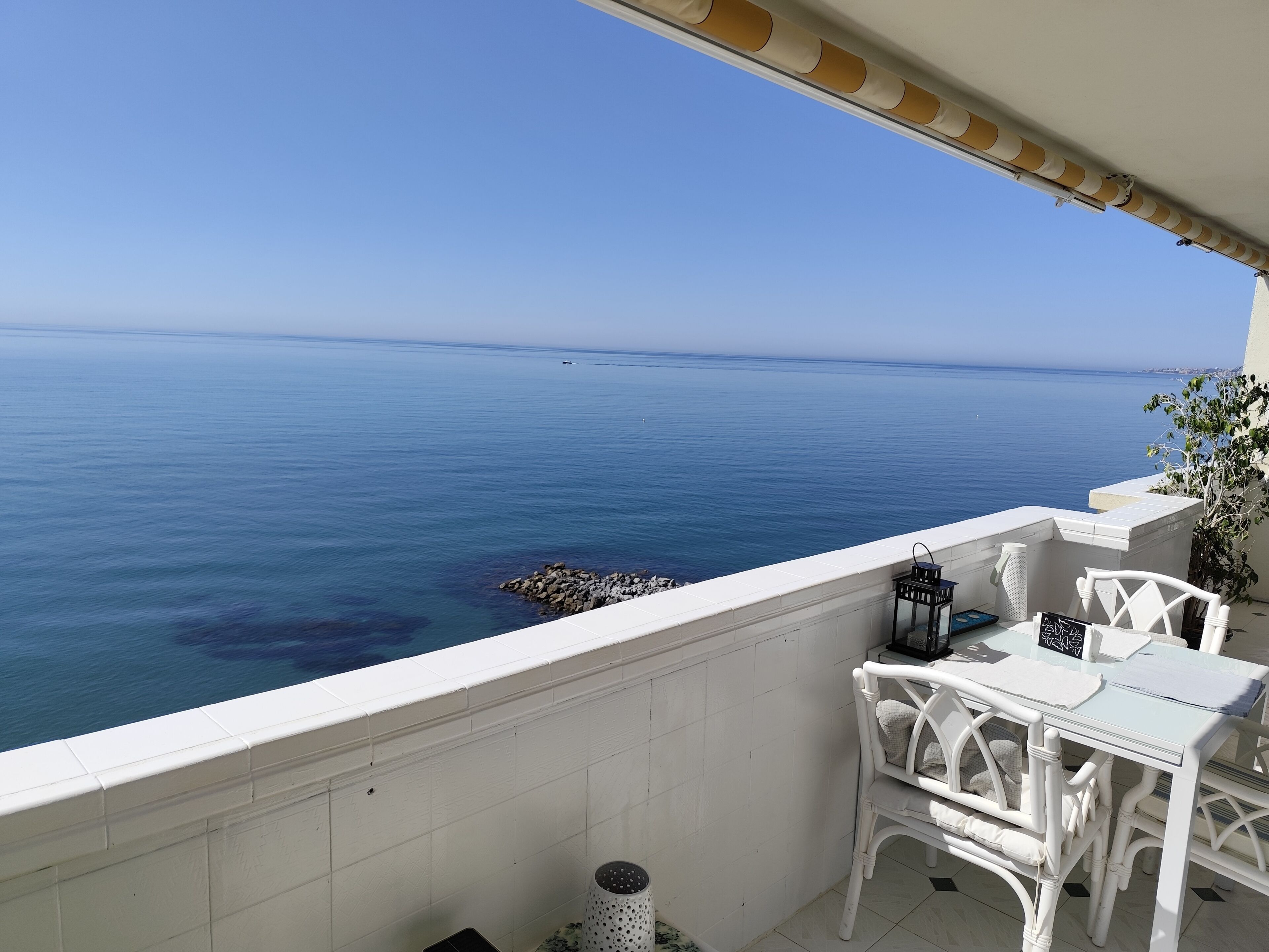 A balcony with a view of the sea, white outdoor furniture, and a tiled floor.