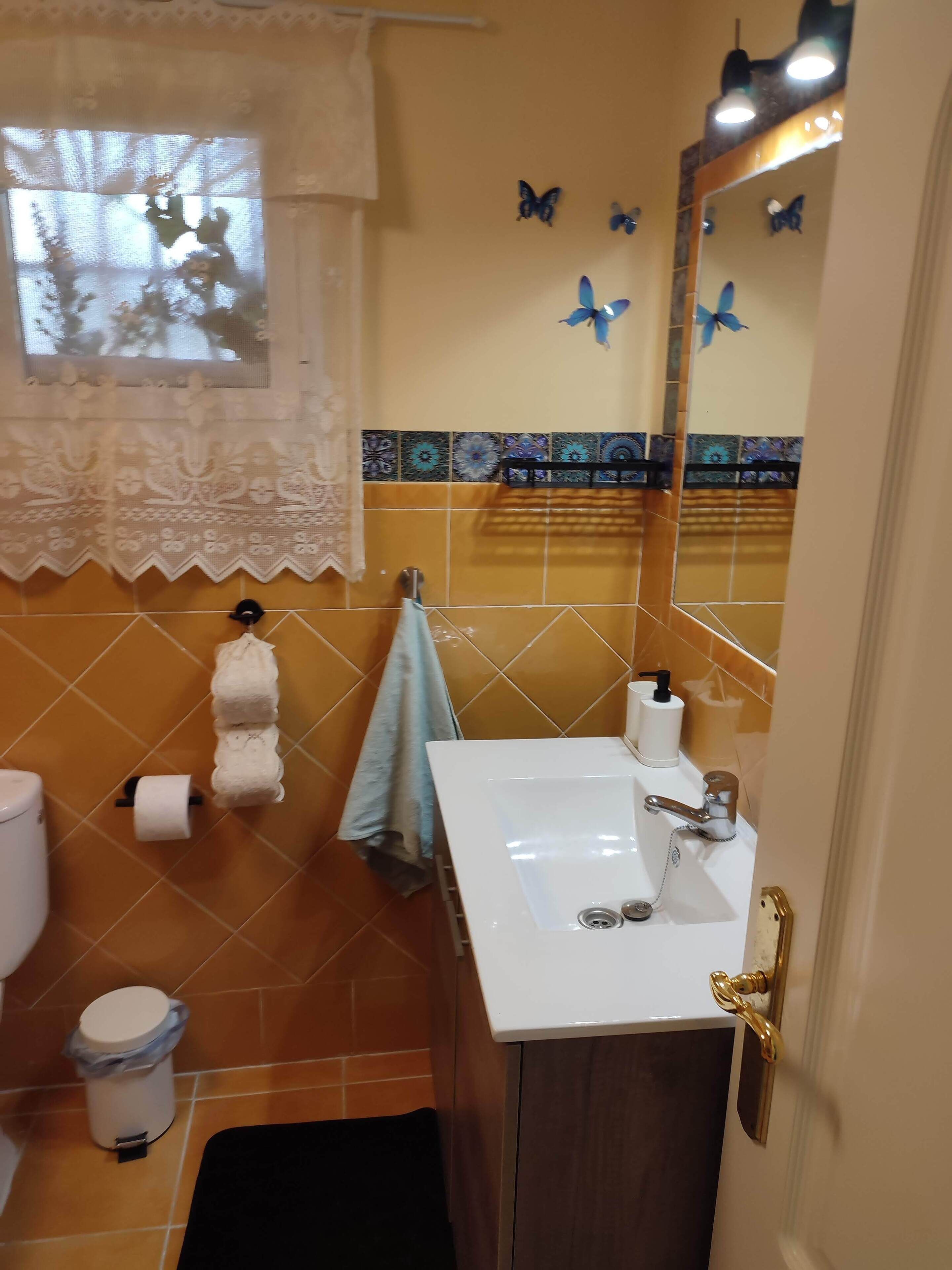 Een badkamer met een wit wastafelmeubel, toilet, prullenbak en raam met kantgordijnen.