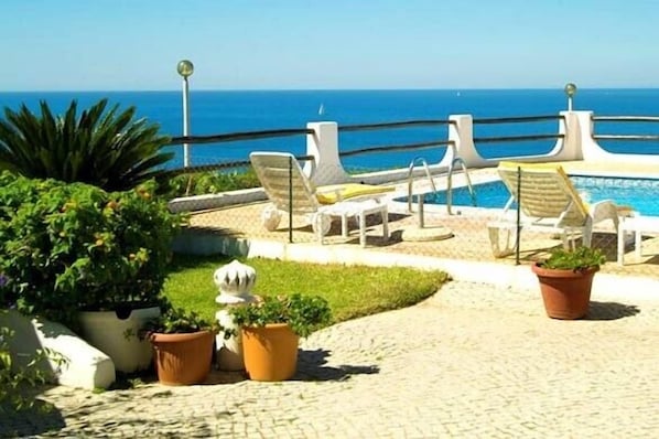 Zona de piscina con sillones de descanso y plantas en macetas, con vistas a un mar azul y despejado.