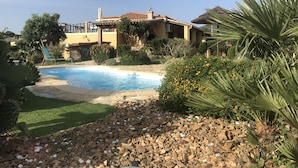 Une piscine entourée d’un ciel dégagé, une maison avec un toit en tuiles et une végétation luxuriante.