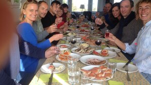 Un grupo de personas sentadas alrededor de una mesa con diversos platos y bebidas, en una sala con ventanas y un candelabro.