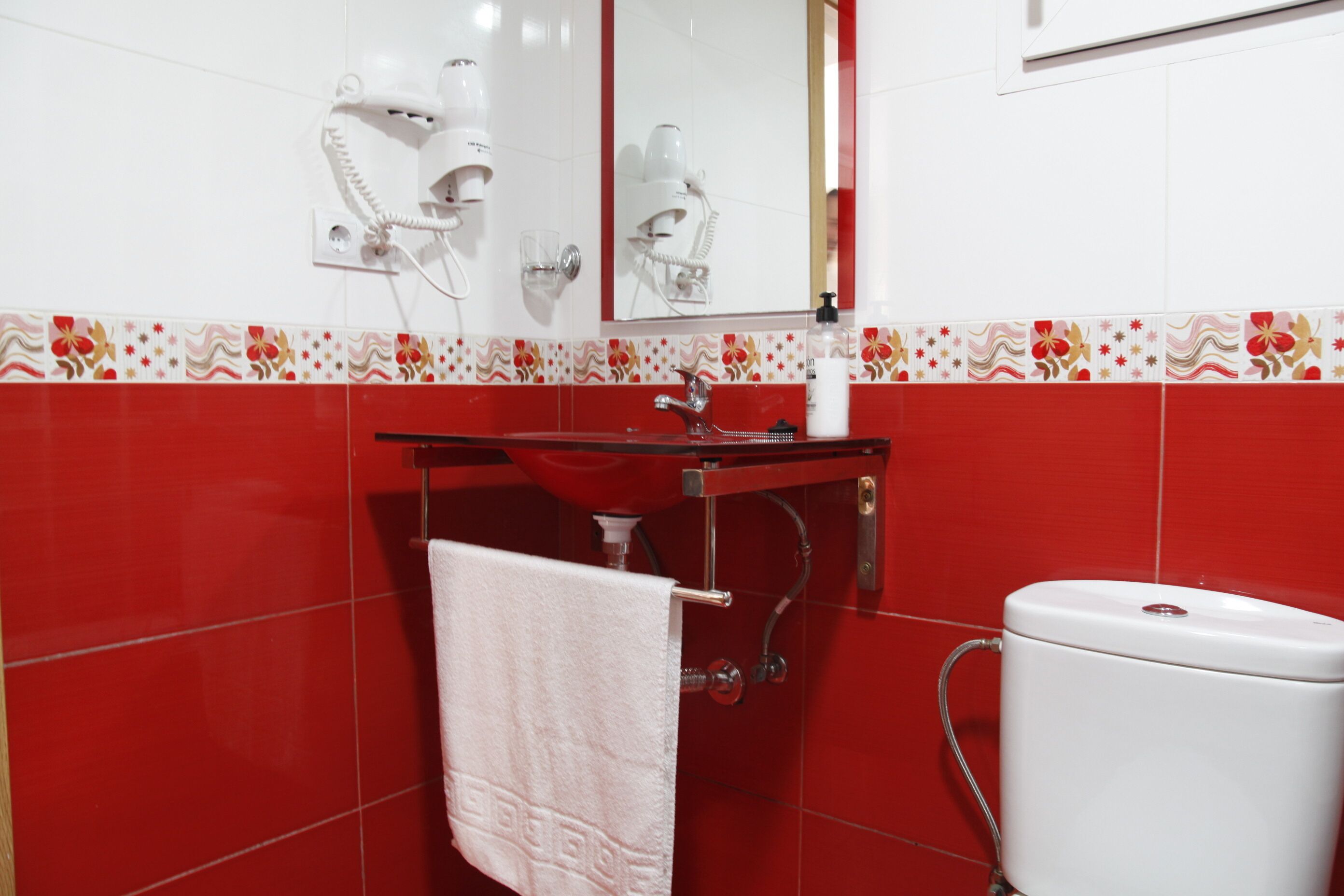 Une salle de bain avec un mur recouvert de carreaux rouges, un lavabo blanc, un miroir, un sèche-cheveux et des toilettes.