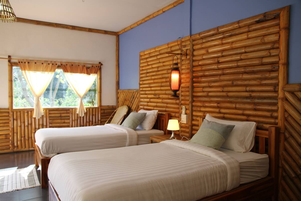 A room with two beds, a bamboo headboard, a window with curtains, and a nightstand with a lamp.
