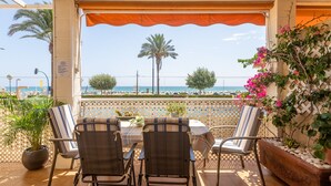 Une terrasse couverte, dotée d’une table et de chaises, qui offre une vue sur une plage bordée de palmiers et un ciel dégagé.