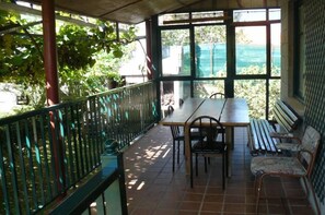 Zona pranzo all'aperto coperta, con tavolo e sedie, una panca e vista su piante e edifici attraverso ampie finestre.