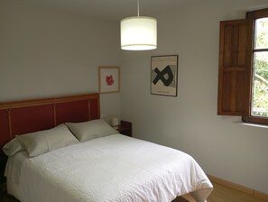 A bed with a white bedspread, a wooden headboard, a nightstand with a lamp, a framed artwork, and a window with wooden shutters.