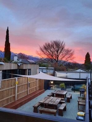 Une terrasse sur le toit, avec des tables et des bancs en bois, un auvent blanc et une vue sur une chaîne de montagnes au coucher du soleil.
