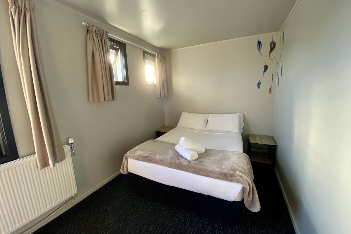 Habitación de hotel con cama, mesita de noche, ventana con cortinas, radiador y adornos en la pared.