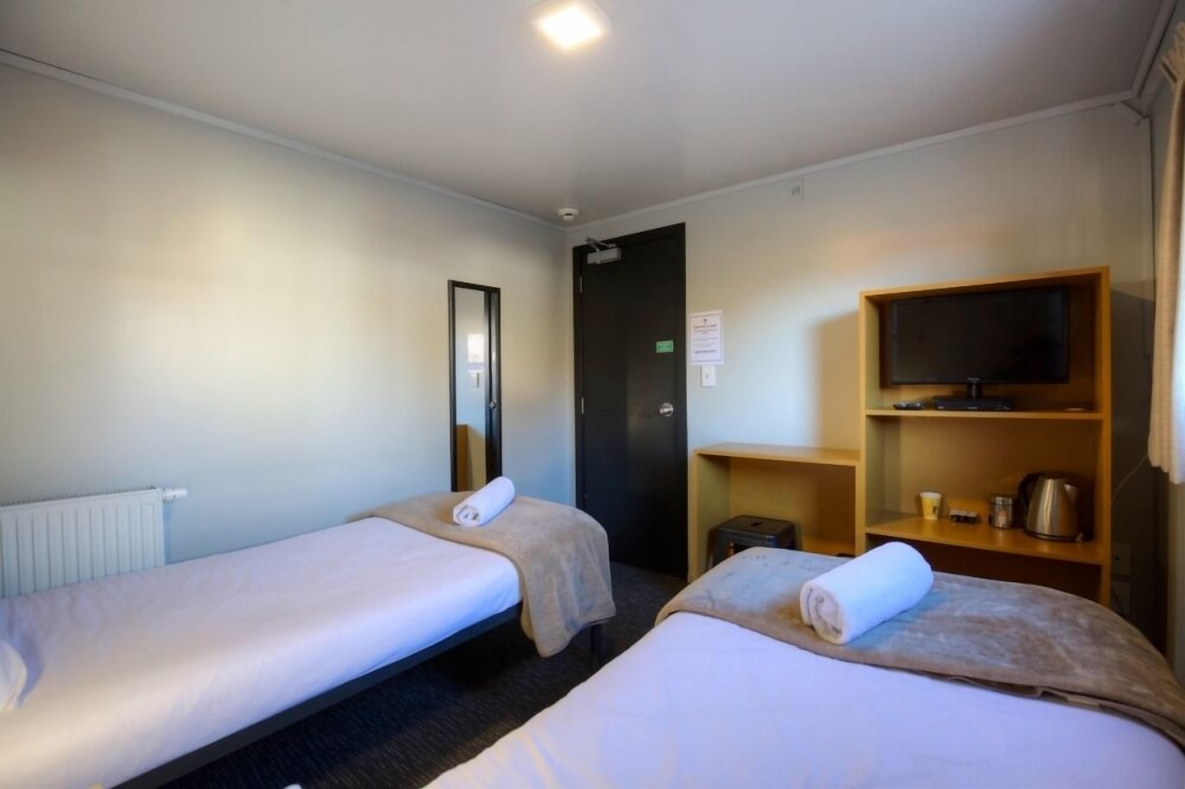 Une chambre d’hôtel avec deux lits simples, un téléviseur posé sur une étagère en bois, un radiateur et une porte avec une pancarte.
