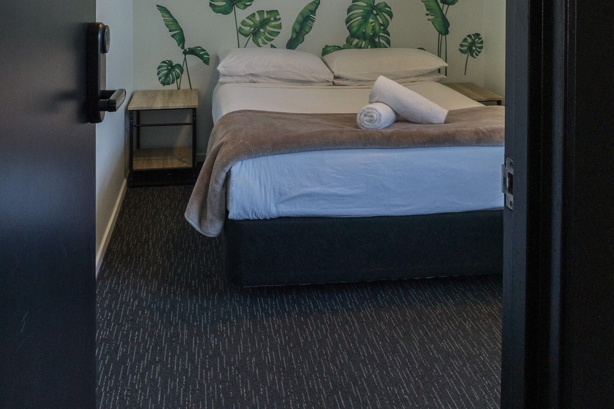 Una habitación de hotel con cama, mesita de noche, lámpara de pared y puerta con número.