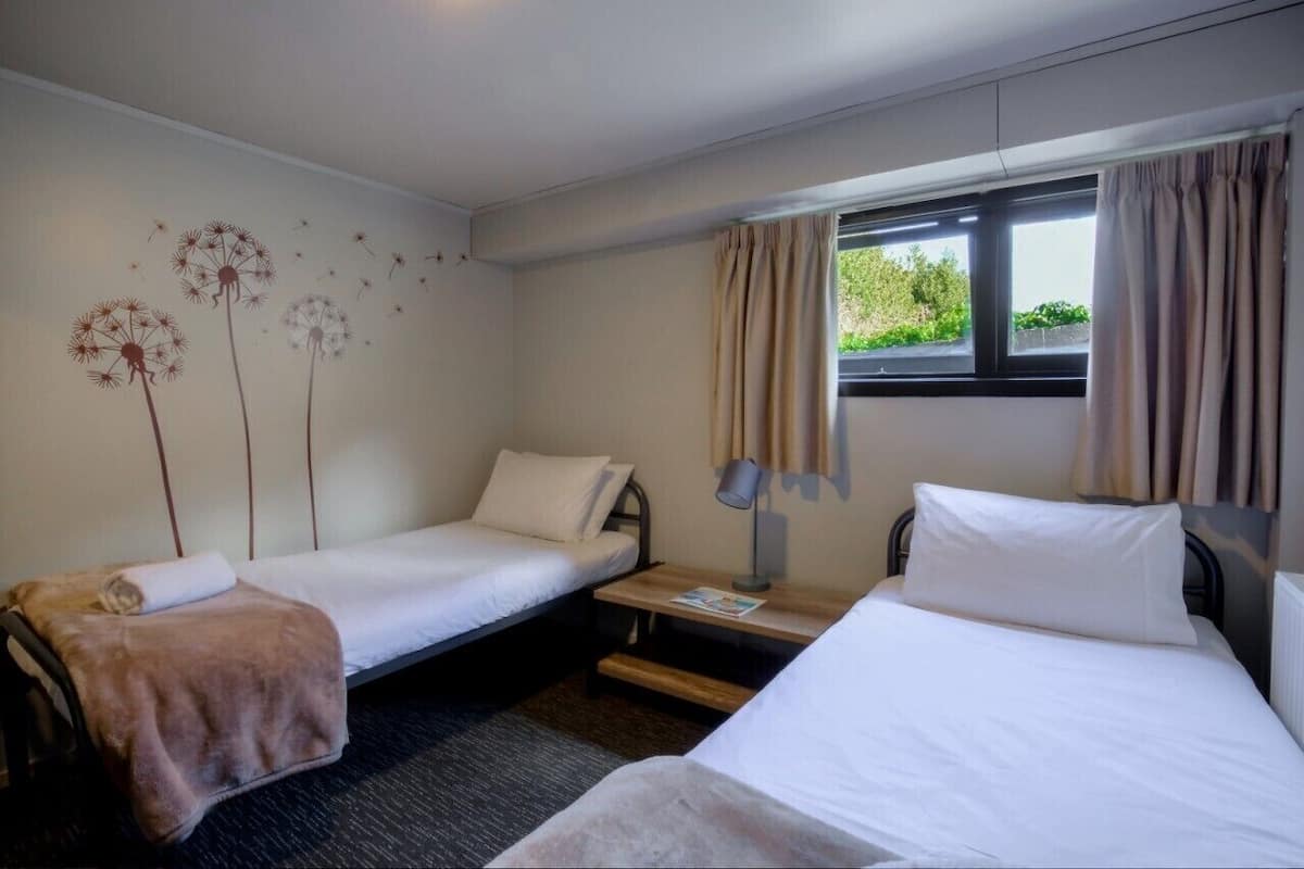 Habitación de hotel con dos camas individuales, una mesita con lámpara, una ventana con cortinas y un cuadro con diente de león.