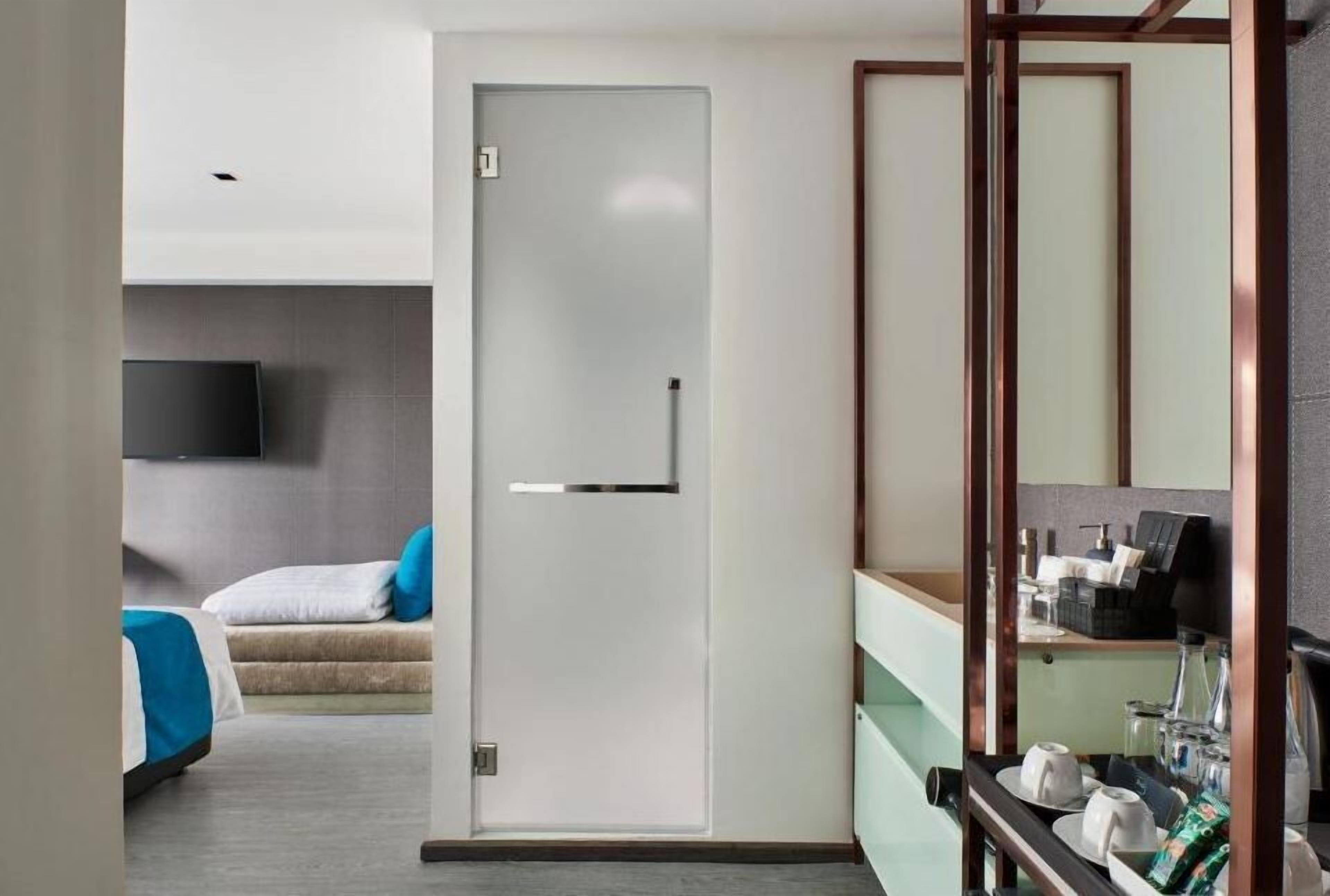 Une chambre d’hôtel moderne au design minimaliste, comprenant un lit avec une literie bleue et blanche, un petit coin salon, un téléviseur à écran plat fixé au mur, et une salle de bain avec une douche en verre et un meuble-lavabo avec des articles de toilette.
