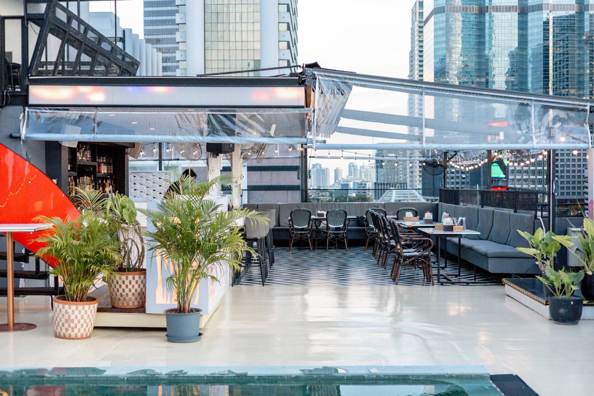 A rooftop bar with seating, potted plants, and a view of the city skyline.