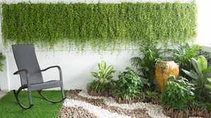 Un jardín con una silla de mimbre negro, diversas plantas en macetas y un jarrón decorativo.