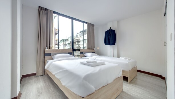Habitación de hotel con dos camas, un ventanal con cortinas y vistas a edificios, además de un perchero con una chaqueta.