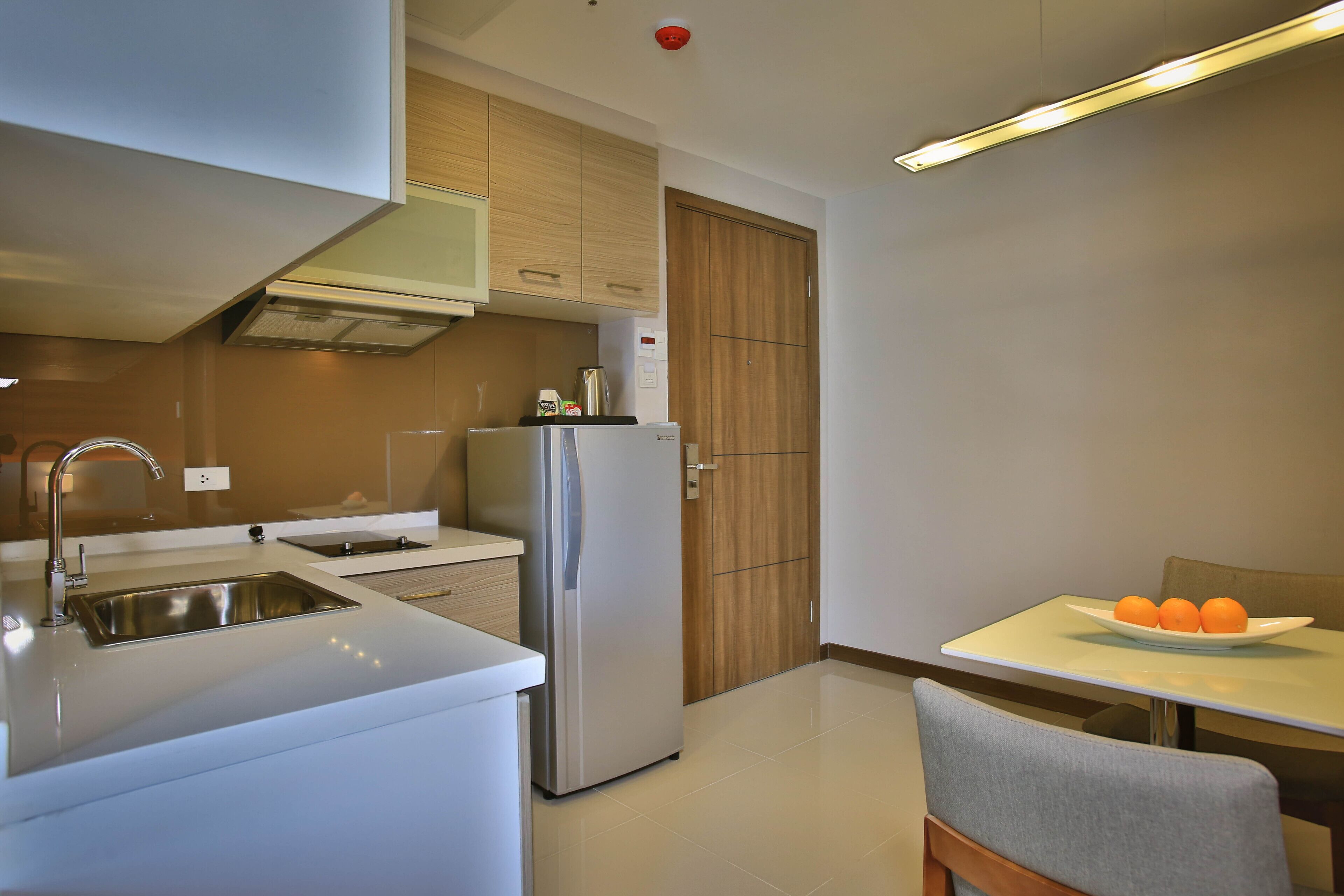 Una cocina moderna con un lavamanos de acero inoxidable, un refrigerador y un mueble de madera. Hay una mesa con un plato de naranjas y una silla.