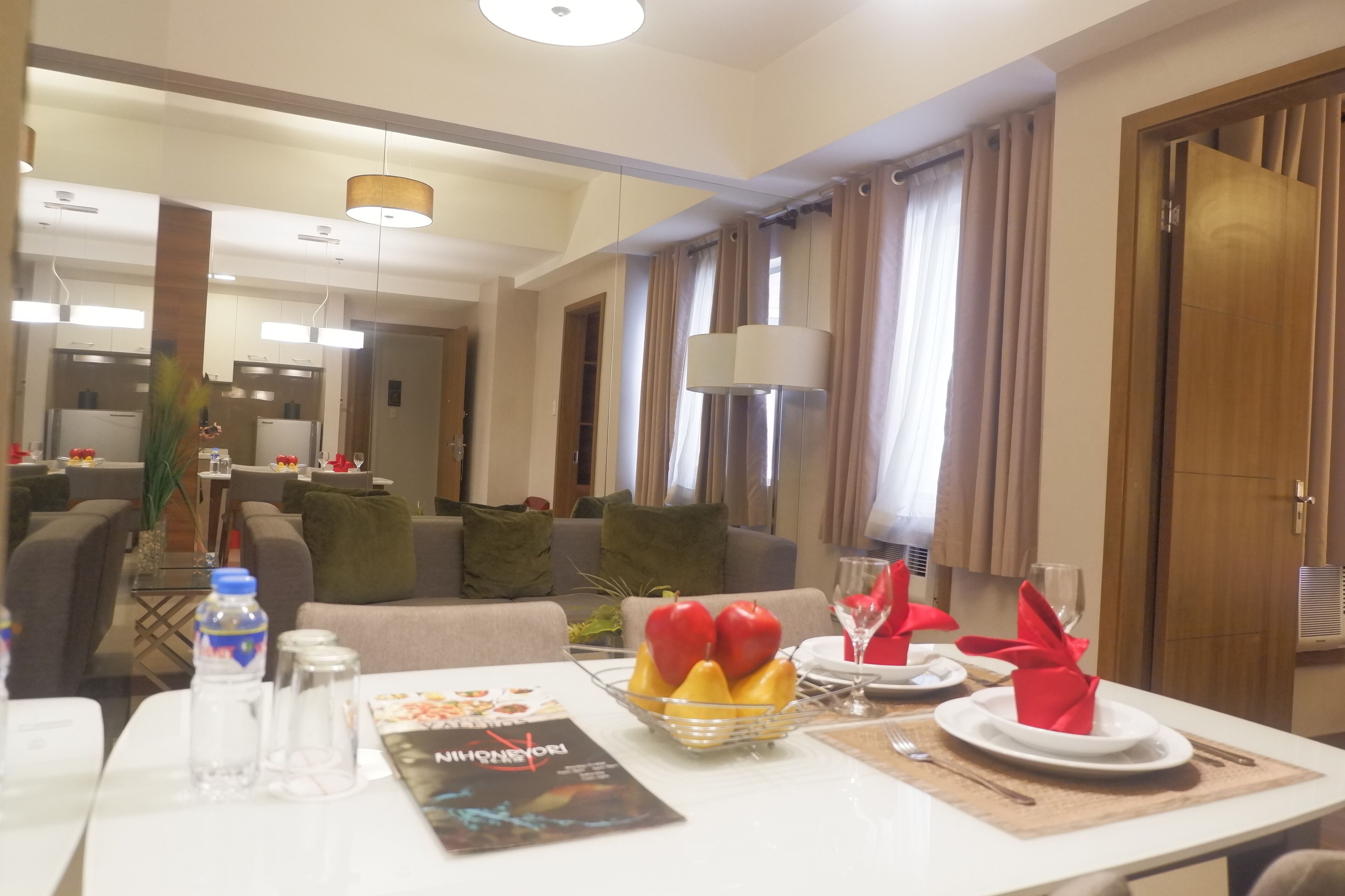 A hotel room with a dining table set for two, a fruit bowl, and a magazine. There is a sofa and a small table with a plant in the background.