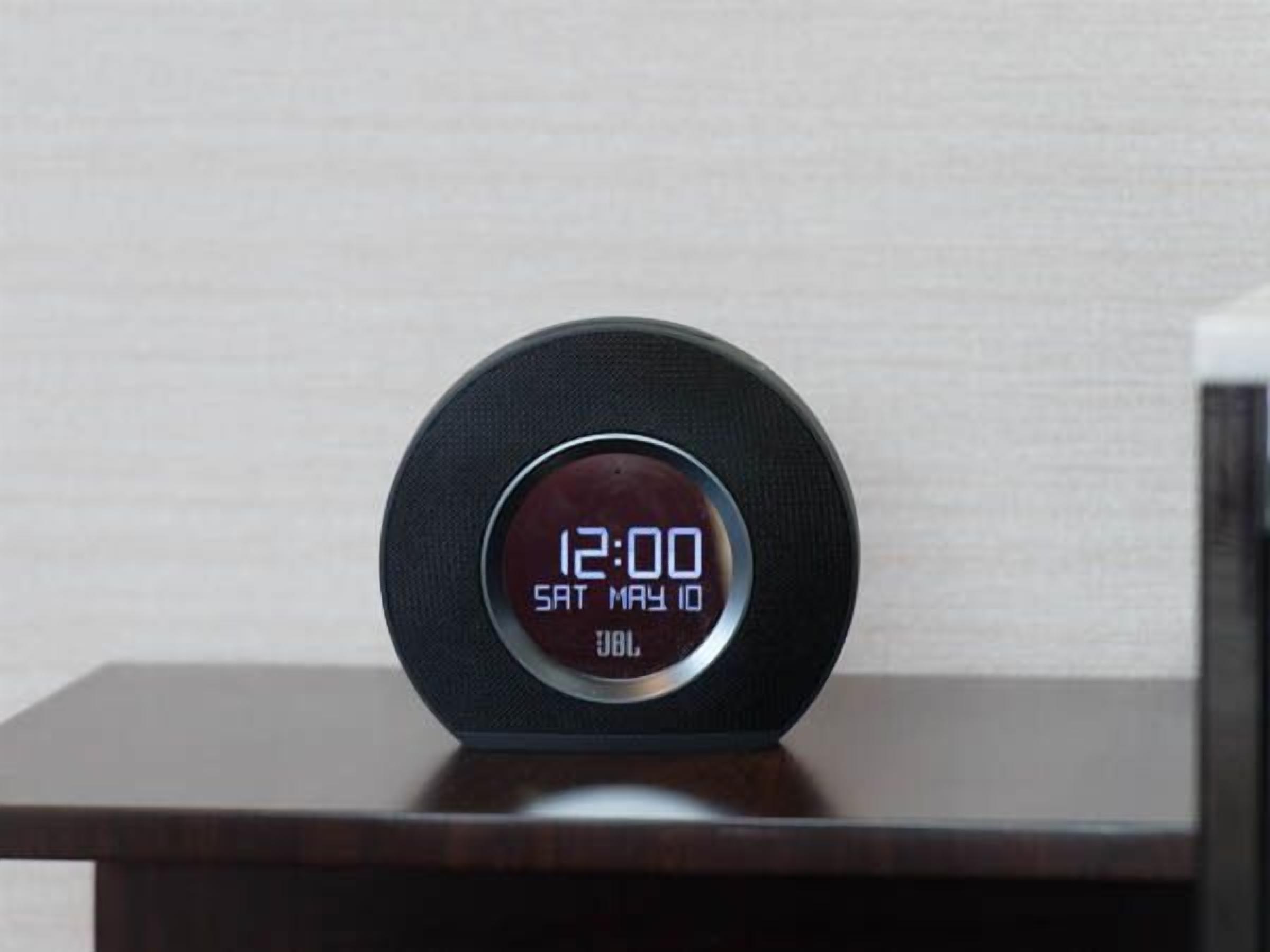 Un reloj digital muestra las 12:00 sobre una mesa de madera oscura frente a una pared lisa.