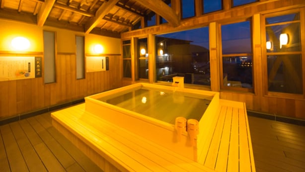 A wooden spa with a hot tub, wooden beams, and large windows overlooking a cityscape at dusk. - LiVEMAX RESORT AKIMIYAJIMA (Hatsukaichi)