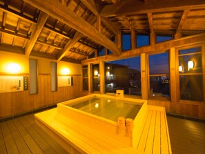 A wooden spa with a hot tub, wooden beams, and large windows overlooking a cityscape at dusk. - LiVEMAX RESORT AKIMIYAJIMA (Hatsukaichi)
