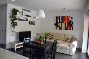 A modern living room with a dining table, chairs, a sofa, a TV, and a colorful abstract painting on the wall.