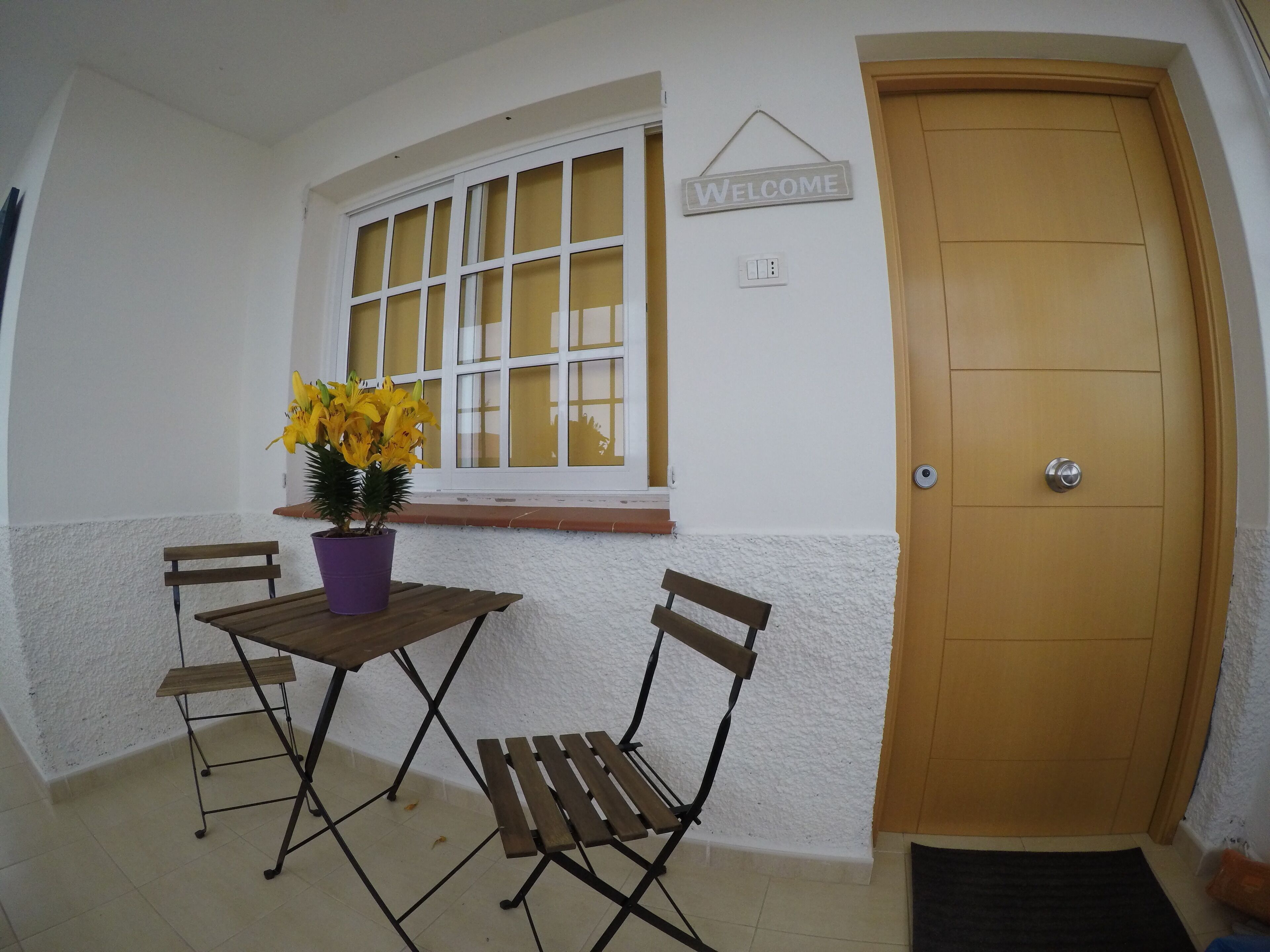 Una habitación con puerta de madera, un cartel de bienvenida, una ventana con cortinas amarillas, una pequeña mesa con sillas y una planta en maceta.