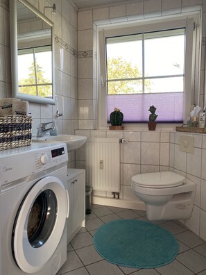 A modern bathroom with a washing machine, sink, toilet, and a window with a view of trees.