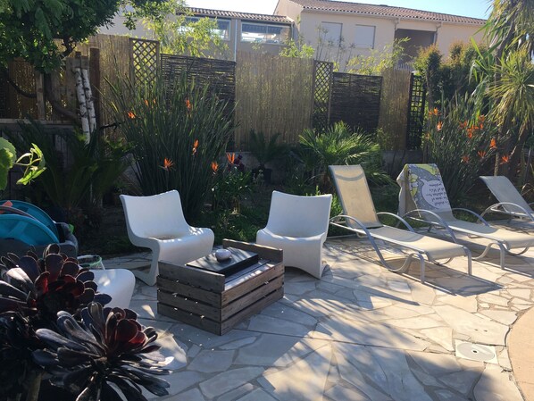 An outdoor patio with white lounge chairs, a wooden table, and a variety of plants.