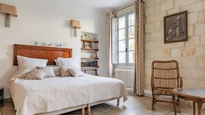 Un dormitorio con cama, mesitas de noche, una silla, una estantería y una ventana con cortinas.