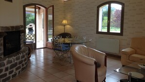 A living room with a stone fireplace, a glass coffee table, a beige armchair, and a view of a garden through large windows. - Spacious villa, private, secure swimming pool Jacuzzi on large, enclosed grounds (Sampzon)
