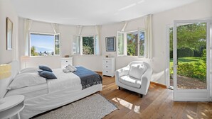 Un dormitorio con cama, mesitas de noche, una silla y ventanas amplias con cortinas.