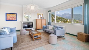 Una sala de estar moderna con sofá, mesa de centro y televisor, ventanas amplias con vistas a la montaña y un aparador de madera.