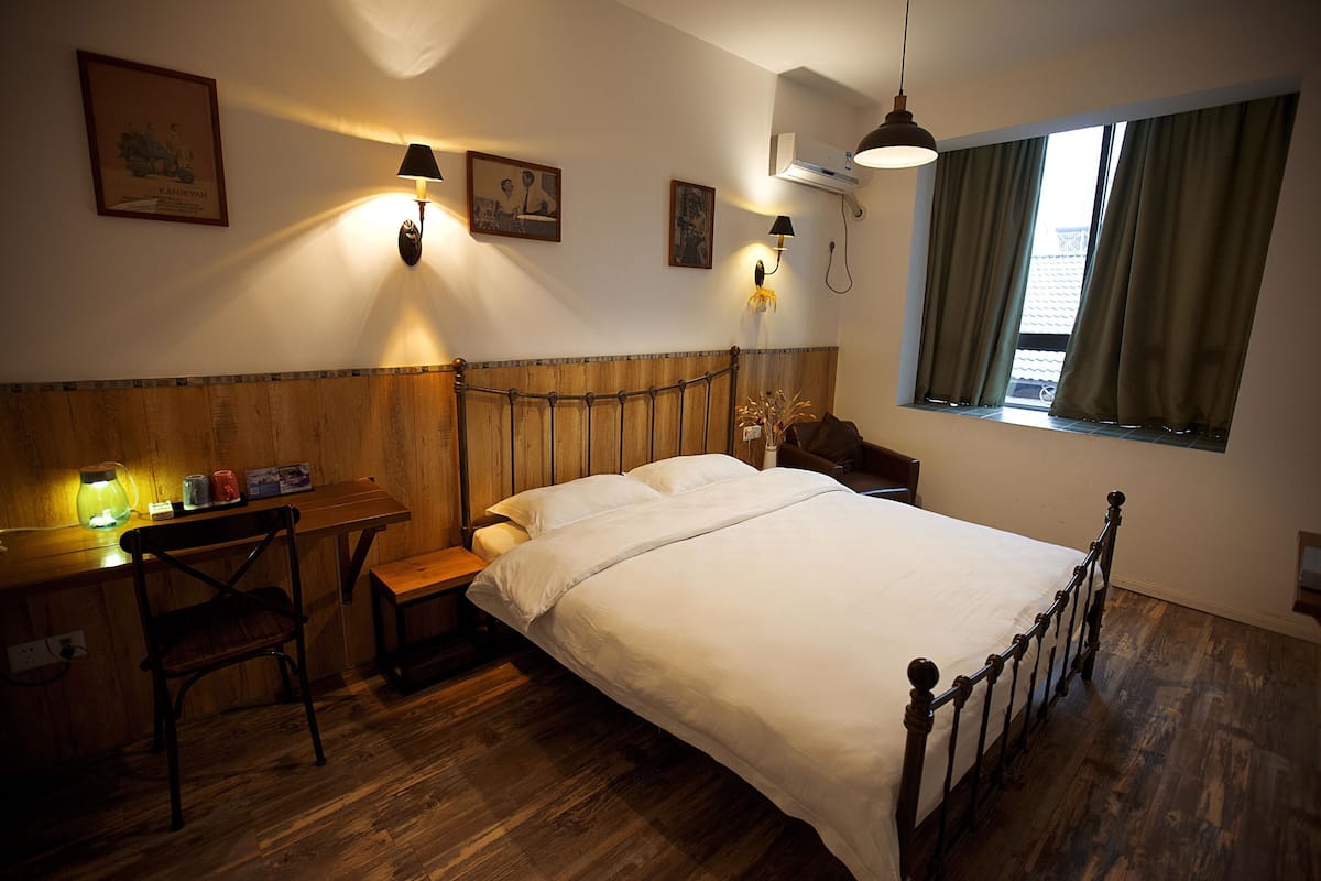 A bedroom with a wooden headboard, a bed with white linens, a desk with a chair, and a window with curtains.