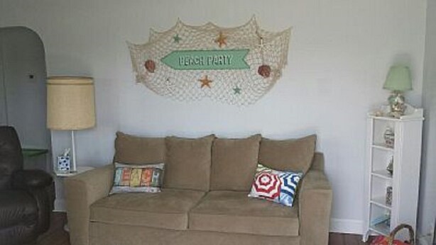 A living room with a beige sofa, two patterned throw pillows, a wall decoration, a lamp, and a small shelf with decorative items.