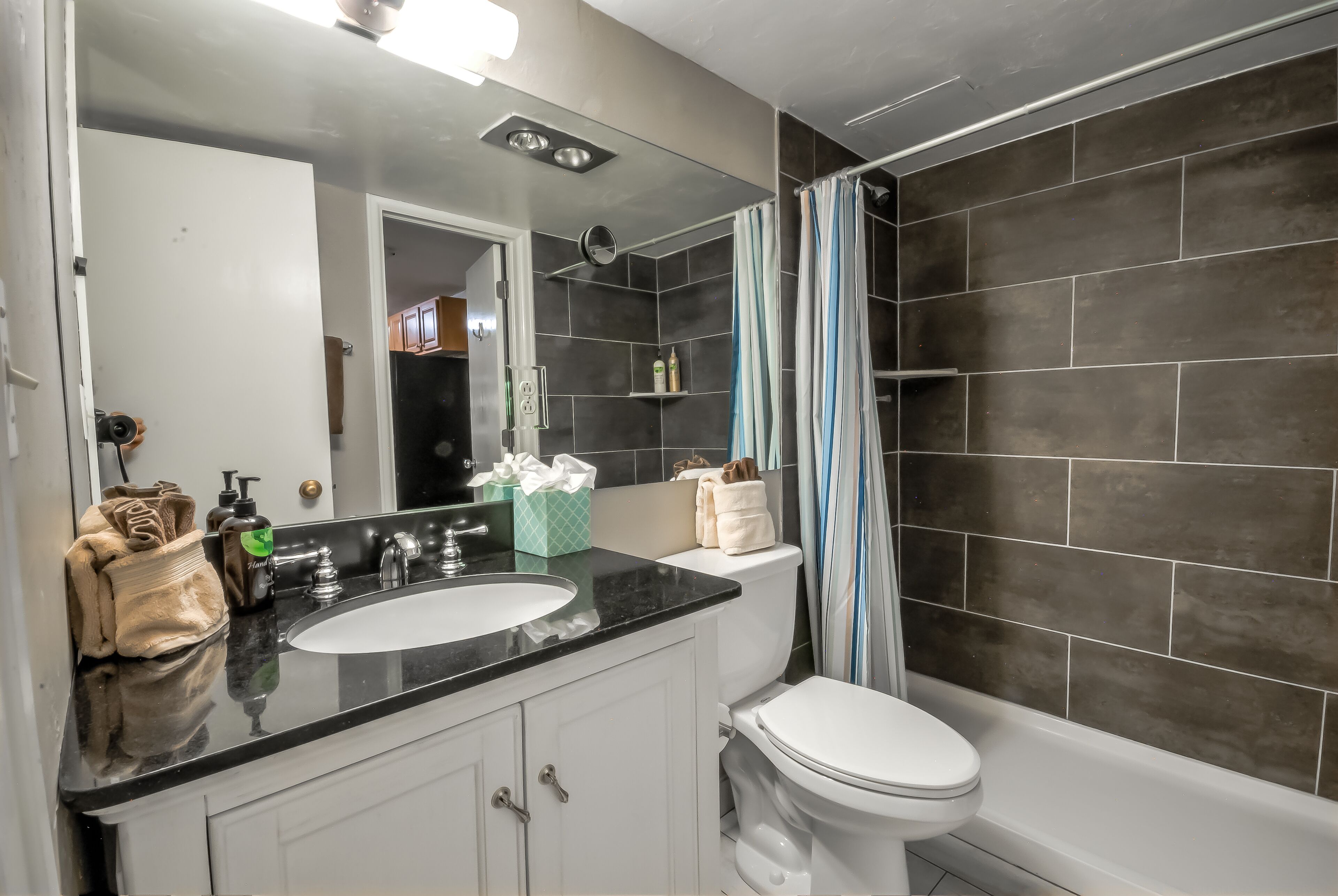 A modern bathroom with a black countertop sink, white cabinets, a white toilet, and dark tiled walls.