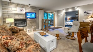 Un salon moderne avec un mur en pierre, un téléviseur à écran plat, une cheminée, un canapé, une table basse, un fauteuil et un lit donnant sur la forêt.