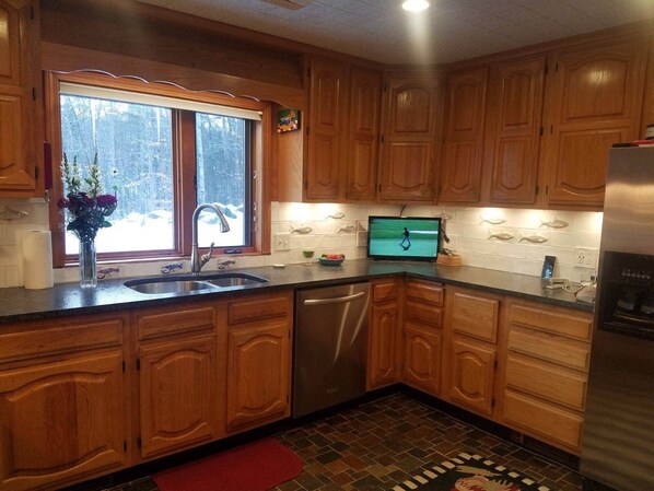 A kitchen with wooden cabinets, a stainless steel refrigerator, a dishwasher, and a window with a view of trees.