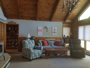 A cozy living room with a wooden ceiling, a fireplace, a bookshelf, a sofa, a coffee table, and a window with blinds. - Your Lakeside Paradise: A/C, Private Dock, Kayaks & Rowboat on Dexter Pond! (Wayne)