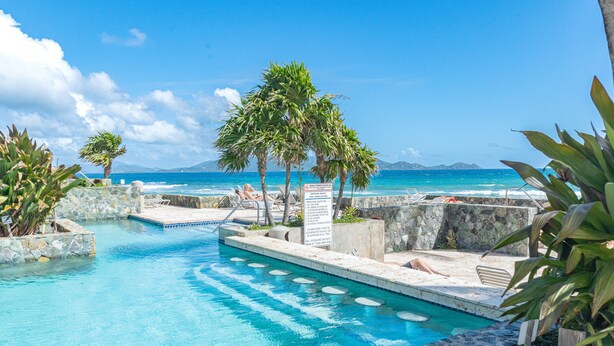 A pool area with clear blue water, surrounded by stone walls and palm trees, overlooking a beach and ocean. - Sea Worthy ~ Breezy & beachfront, steps to the water with views of St John (St. Thomas)