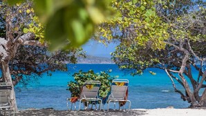 A beach with two lounge chairs and a small table, surrounded by trees and overlooking a clear blue sea. - Sea Worthy ~ Breezy & beachfront, steps to the water with views of St John (St. Thomas)