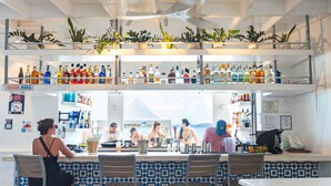 A bar area with multiple shelves of bottles, a counter with a 'ORDER HERE' sign, and a view of the sea. - Sea Worthy ~ Breezy & beachfront, steps to the water with views of St John (St. Thomas)
