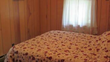 Ein Schlafzimmer mit Holzwänden, einem Bett mit floralem Überwurf, einem Fenster mit leichtem Vorhang und einem kleinen Tisch mit gemustertem Tischläufer.
