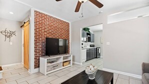 A modern living room with a brick accent wall, a flat-screen TV, a ceiling fan, and a kitchen in the background. - Beach Cottage -optional BIKES - Walk 2 Shops & Fun (Saint Simons Island)