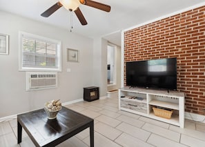 Un salon avec un téléviseur fixé sur un mur en briques, un ventilateur de plafond, une fenêtre avec des stores et une petite table sur laquelle est posé un vase décoratif.