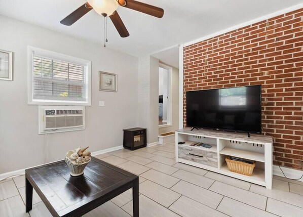 A living room with a TV on a brick wall, a ceiling fan, and a fireplace. - Beach Cottage -optional BIKES - Walk 2 Shops & Fun (Saint Simons Island)
