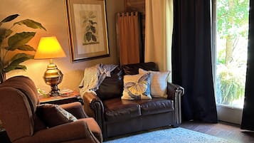 A cozy living room with a brown leather sofa, a patterned rug, a wooden ceiling, and a large window with curtains.