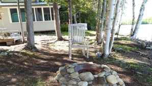 A rustic outdoor setting with a stone fire pit, a wooden bench, and a house in the background. - 10 Steps And You're  In Sebasticook Lake. This Is Where You Go To Unplug (Newport)