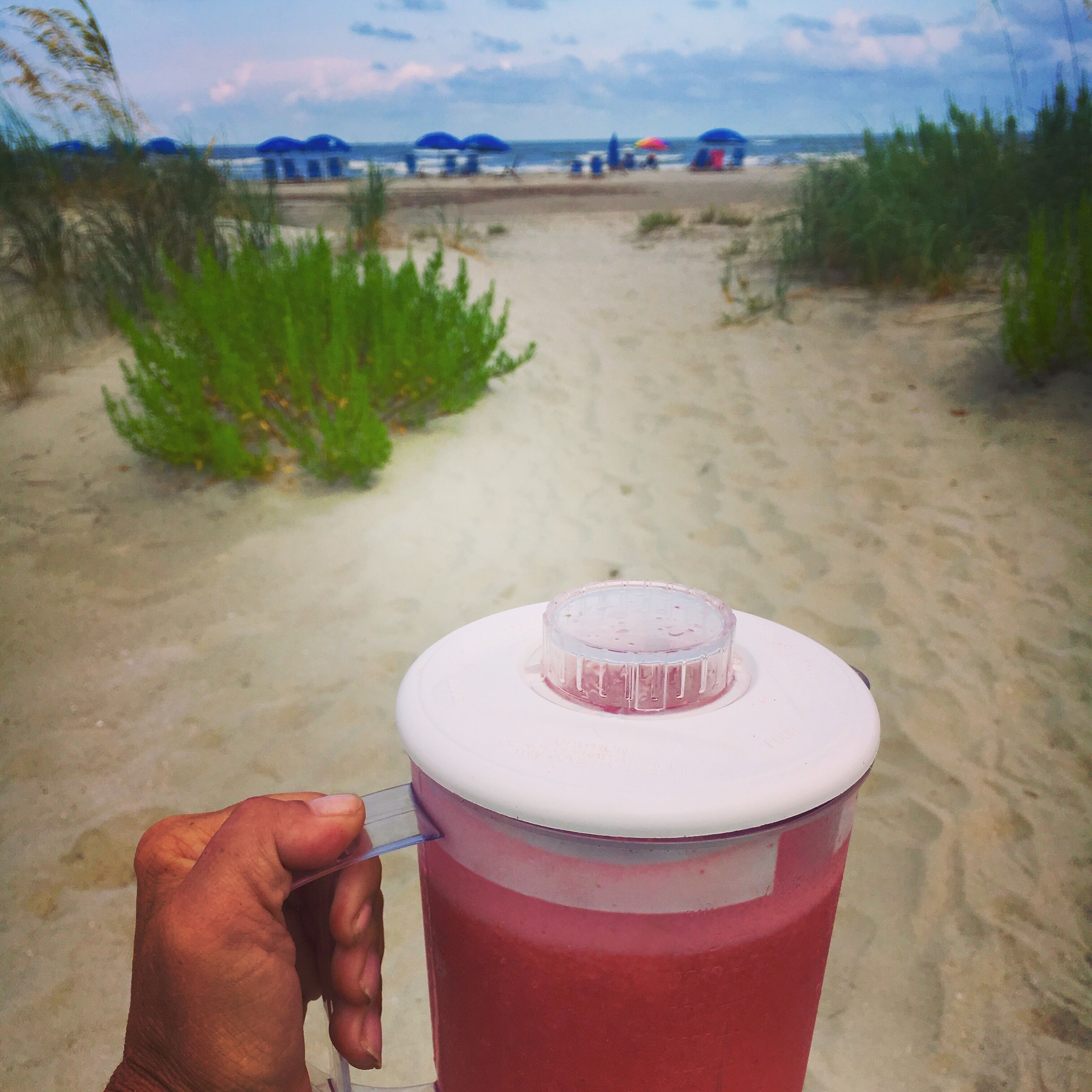 A hand holding a red container with a white lid, placed on a sandy beach with a view of the ocean and beach umbrellas in the background.