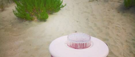A hand holding a red container with a white lid, placed on a sandy beach with a view of the ocean and beach umbrellas in the background.